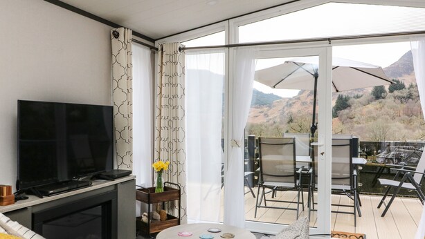 A modern living room with a flat-screen TV, a sofa, a coffee table, and a balcony with outdoor seating. - Red Kite Lodge (Crieff)