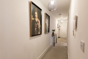 A hallway with framed artwork on the walls, a potted plant on a side table, and a modern pendant light. - The Courtyard (Southampton)