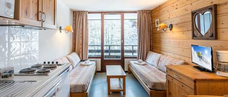 Une chambre confortable de style cabane, avec des murs en bois, une kitchenette, deux lits avec une literie assortie, une télévision et une fenêtre avec des rideaux.