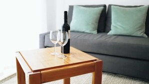 Une table en bois sur laquelle sont posés une bouteille de vin et deux verres, placée devant un canapé gris agrémenté de coussins verts.