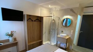 Una habitación de hotel moderna con una cama grande, un armario de madera, una cabina de ducha de vidrio, un espejo redondo, un lavamanos pequeño y un televisor montado en la pared.