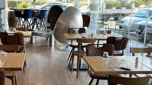 Interior de restaurante con mesas y sillas de madera, bombillas colgantes y grandes ventanales que dan a la calle con coches aparcados.