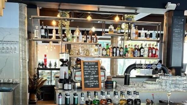 Zona de bar con mostrador de madera, dos taburetes negros y estantes llenos de botellas diversas.