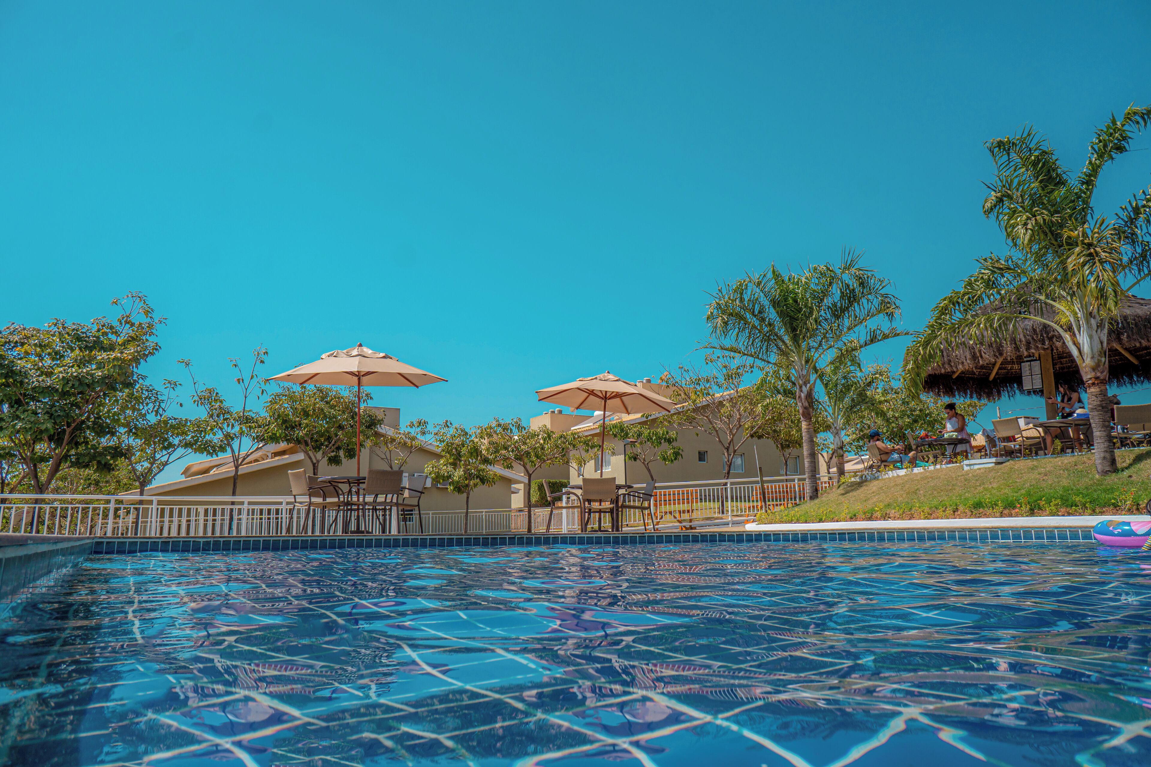 A swimming pool with clear blue water, surrounded by palm trees and umbrellas, and a building with a thatched roof in the background.