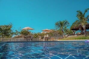 Een zwembad met helderblauw water, omringd door palmbomen en parasols, en een gebouw met rieten dak op de achtergrond.