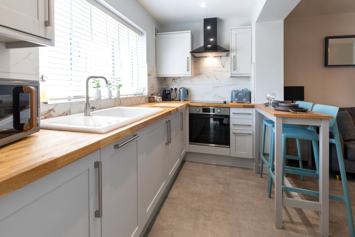 A modern kitchen with white cabinets, a wooden countertop, a sink, a stove, and a dining area with blue chairs.