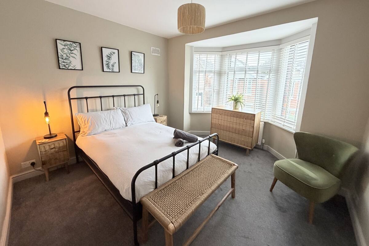 A bedroom with a bed, bedside table, lamp, bench, dresser, chair, and a window with blinds.