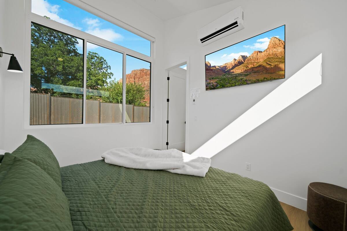 A bedroom with a large window, a bed with green bedding, a mounted TV, and an air conditioning unit.