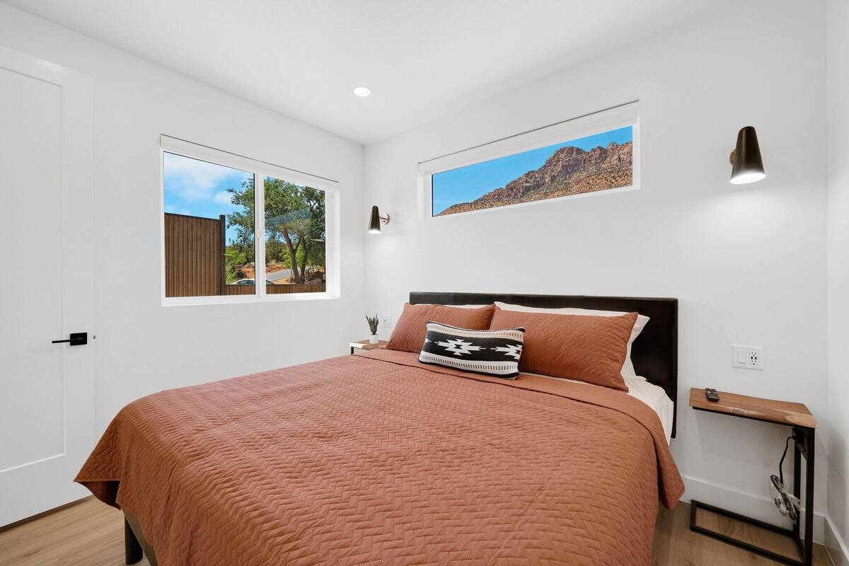 A bedroom with a large bed, a window, a wall-mounted picture, and a bedside table with a lamp.