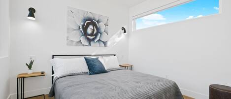 A modern bedroom with a large bed, a bedside table with a plant, a wall-mounted light, and a window with a view of the sky.