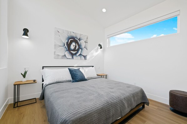 Une chambre moderne avec un grand lit, une table de chevet agrémentée d’une plante, une lampe fixée au mur et une fenêtre donnant sur le ciel.