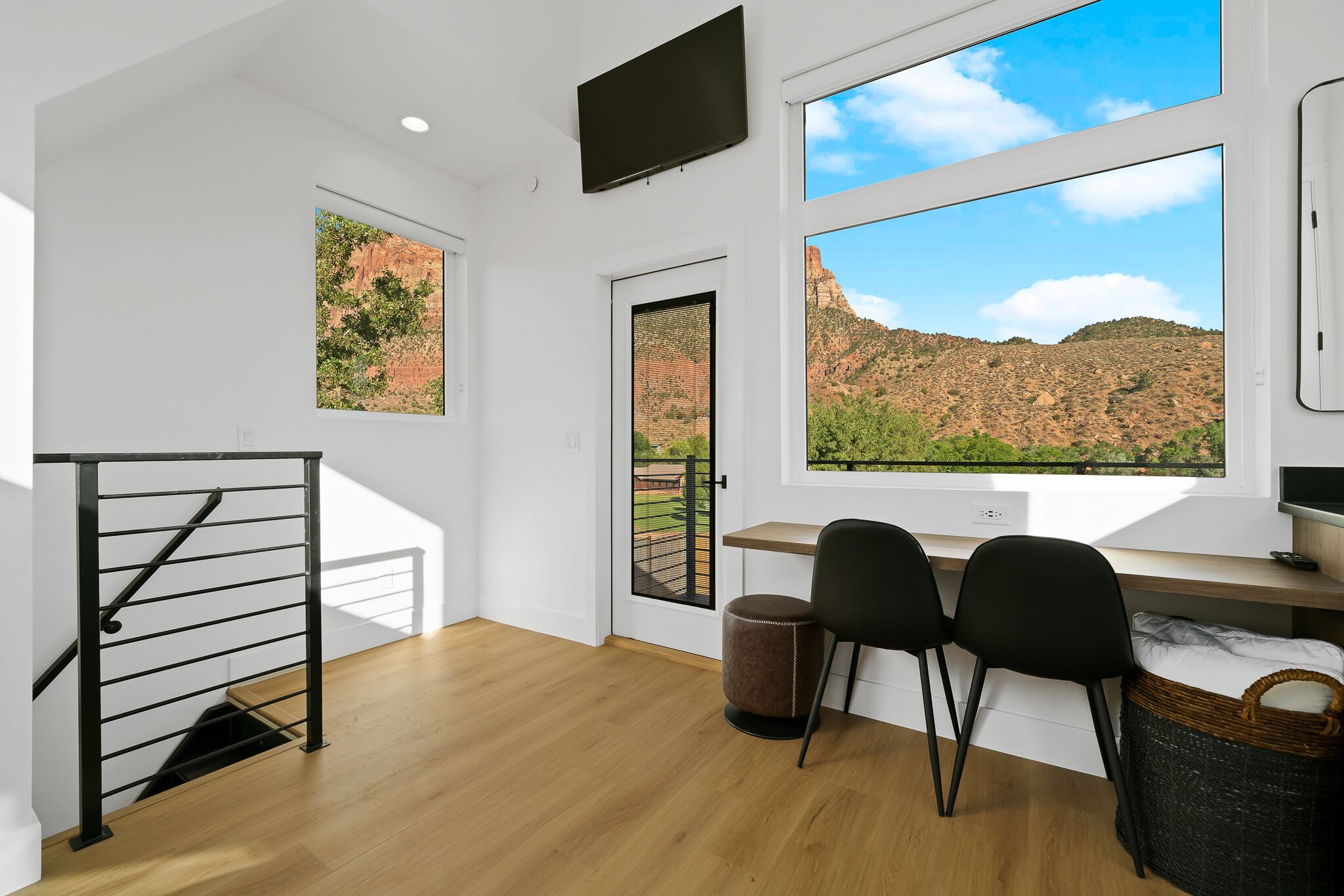 Une chambre moderne avec un plancher en bois, un bureau avec deux chaises, un téléviseur fixé au mur et de grandes fenêtres offrant une vue sur un paysage montagneux.