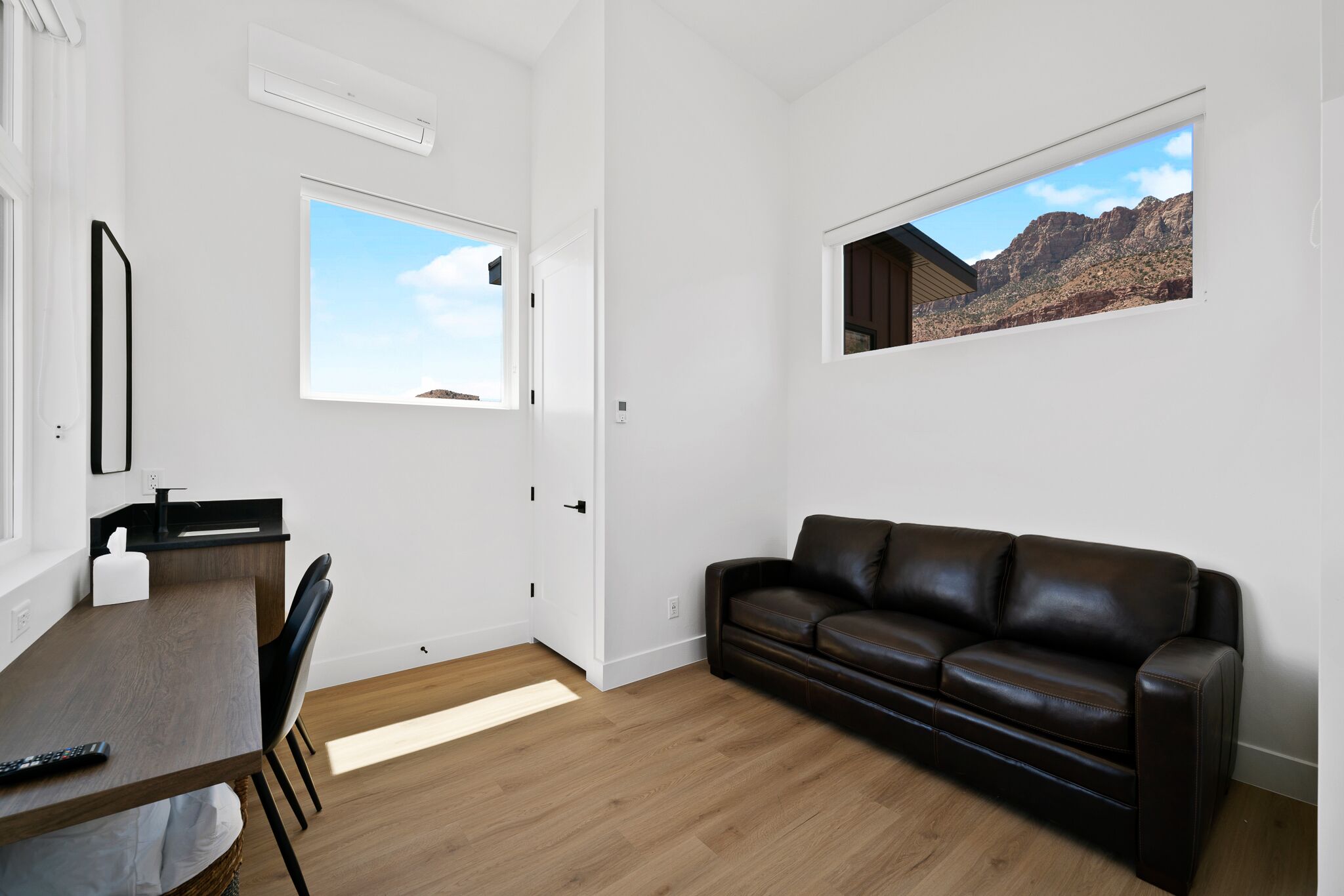 A modern living room with a wooden dining table, a black leather sofa, and a large window with a desert view.