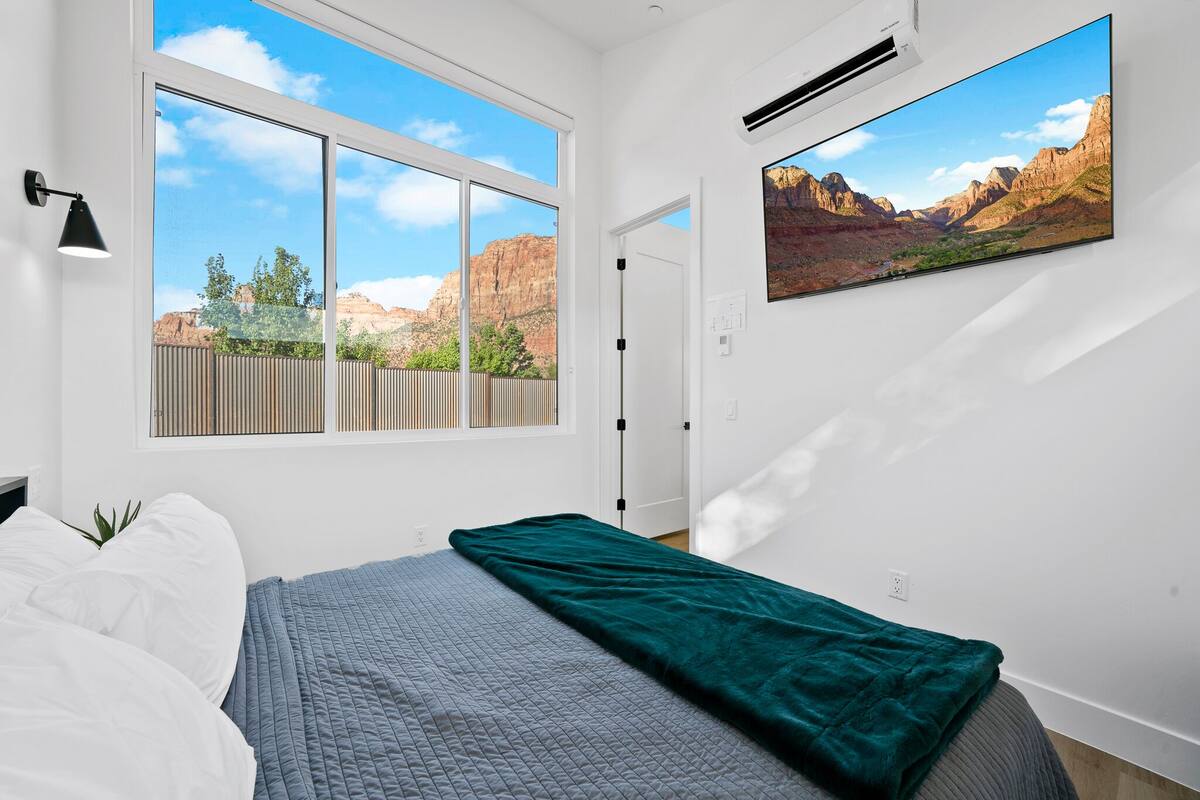 A bedroom with a large window, a bed with a quilt, a green throw blanket, and a wall-mounted TV displaying a mountain landscape.