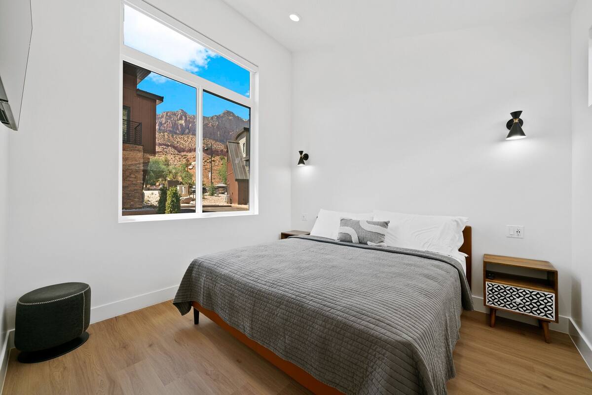 A bedroom with a large window, a bed, a nightstand, and a wall-mounted lamp.