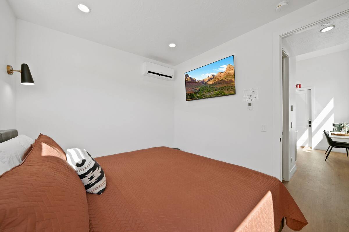 A bedroom with a large bed, a mounted TV, an air conditioning unit, and a door leading to a dining area.