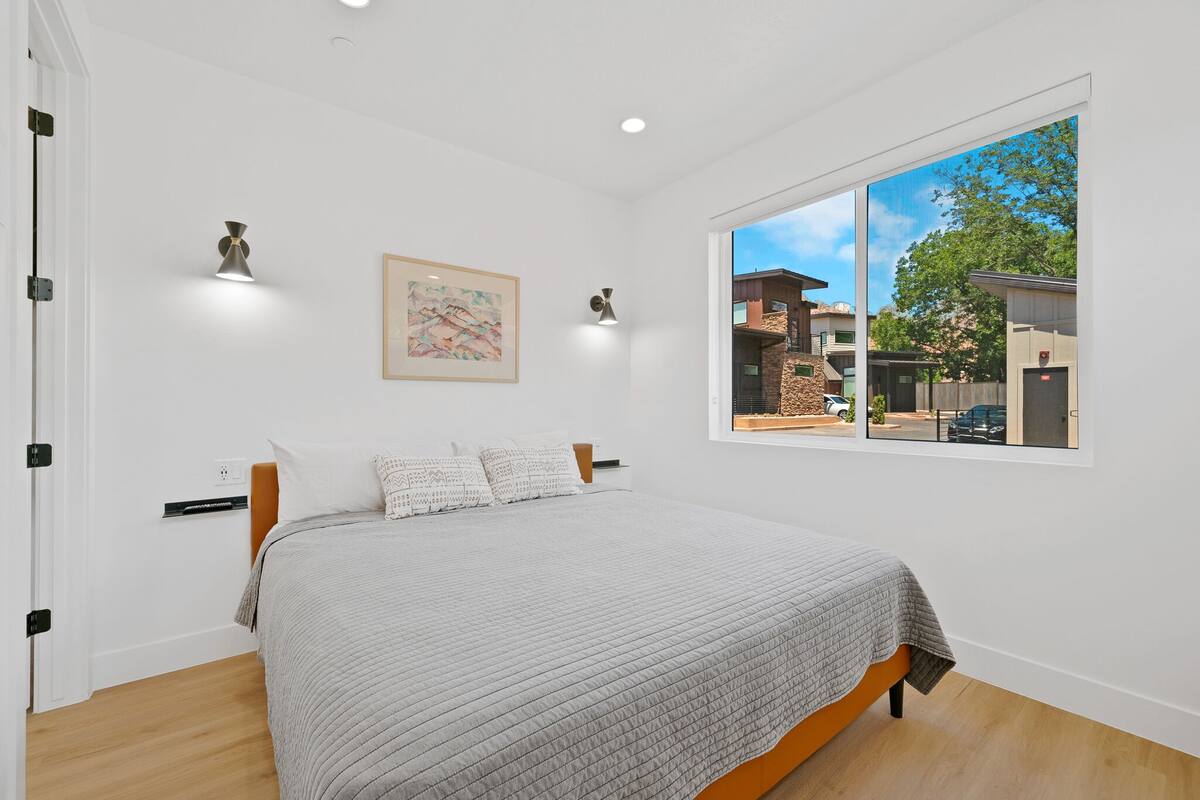 A bedroom with a large bed, a window with a view of buildings, and a wall-mounted light fixture.