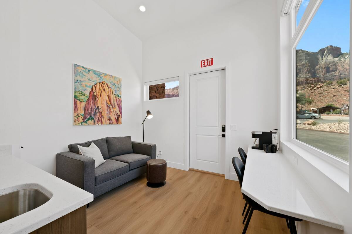A modern room with a gray sofa, a small round ottoman, a white desk, and a window with a view of a mountain landscape.
