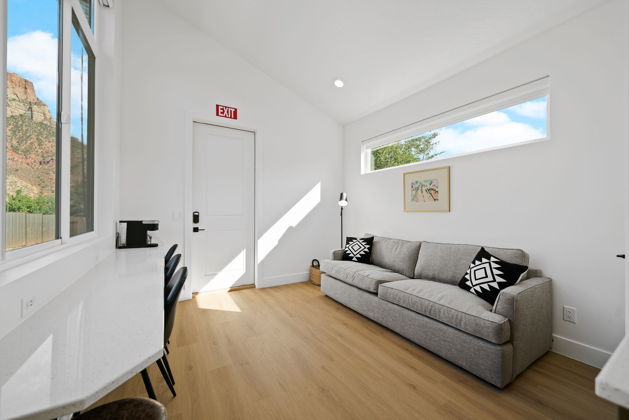 A modern living room with a grey sofa, a dining table with chairs, a white door with an exit sign, and a window with a view of a mountain.