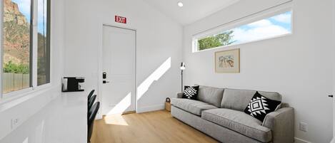 A modern living room with a grey sofa, a dining table with chairs, a white door with an exit sign, and a window with a view of a mountain.