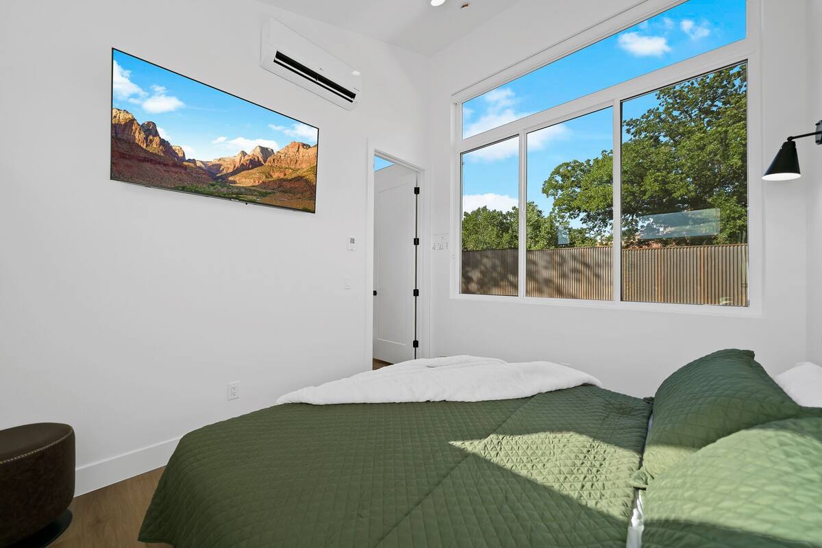 A bedroom with a large bed, a view of the outdoors, and a mounted TV.