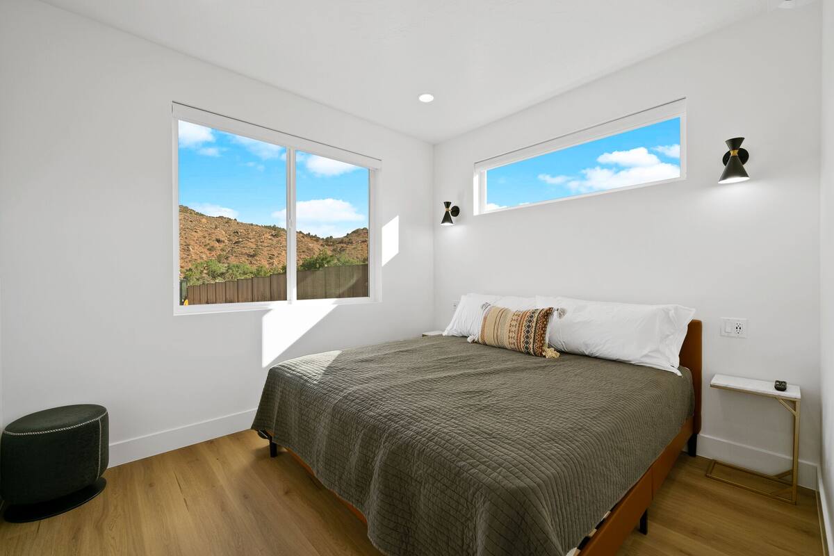 A bedroom with a large bed, two windows, a nightstand, and a view of a hilly landscape.