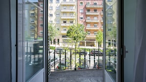 A view from a balcony overlooking a parking area with several cars, a tree, and multi-story buildings in the background. - Home Out 1E - 5 Bedroom Apartament with Parking (Lisboa)