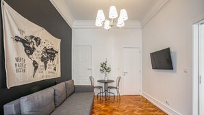 A modern living room with a grey sofa, a wooden herringbone floor, a world map tapestry, a chandelier, a small round table with chairs, and a TV mounted on the wall. - Home Out 1E - 5 Bedroom Apartament with Parking (Lisboa)