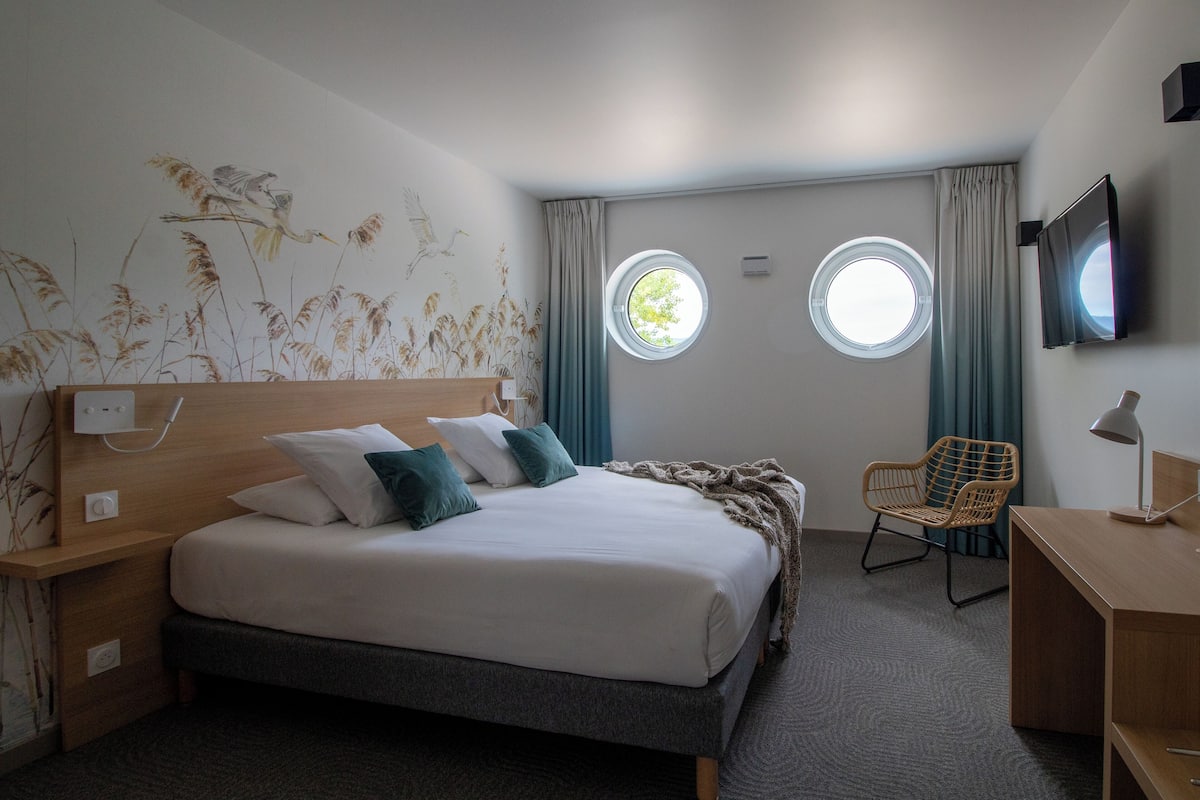 A hotel room with a bed, two windows, a chair, a desk, and a television.