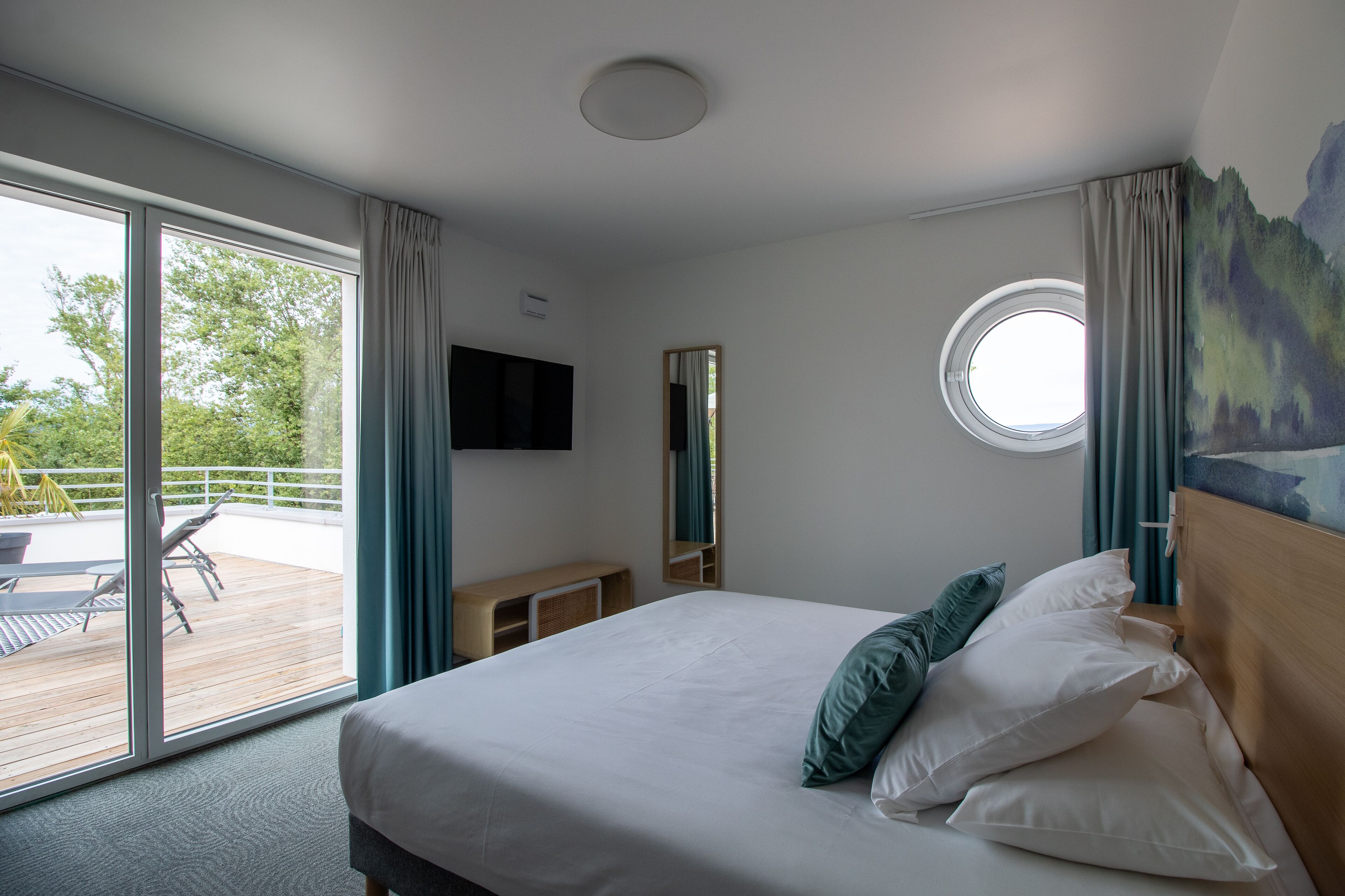 Habitación de hotel con una cama grande, un ventanal redondo, un balcón con mesa y sillas, y un televisor de pared.