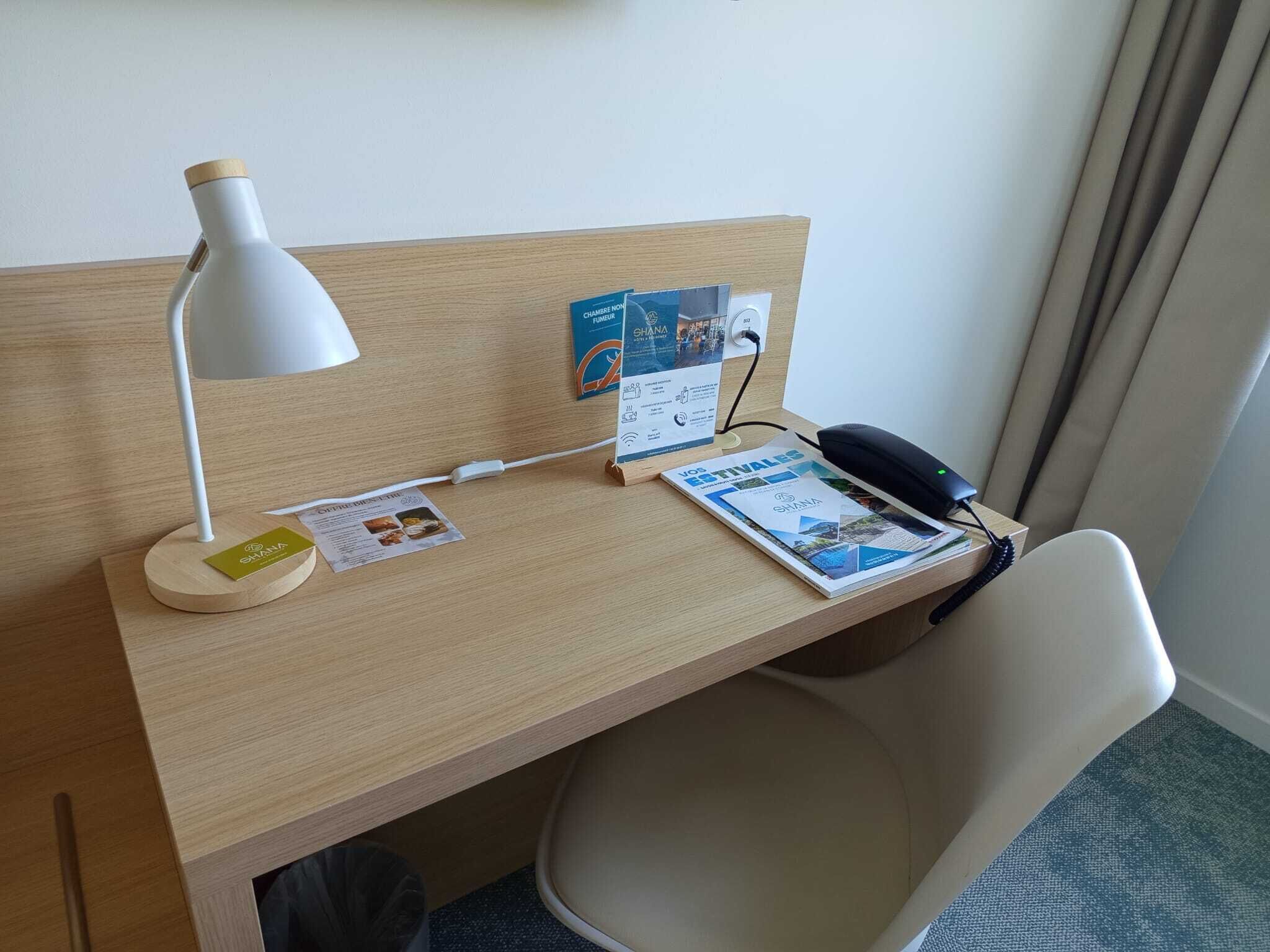 Een hotel bureau met een witte bureaulamp, een zwarte telefoon, een tijdschrift en een klein kaartje op het bureau.