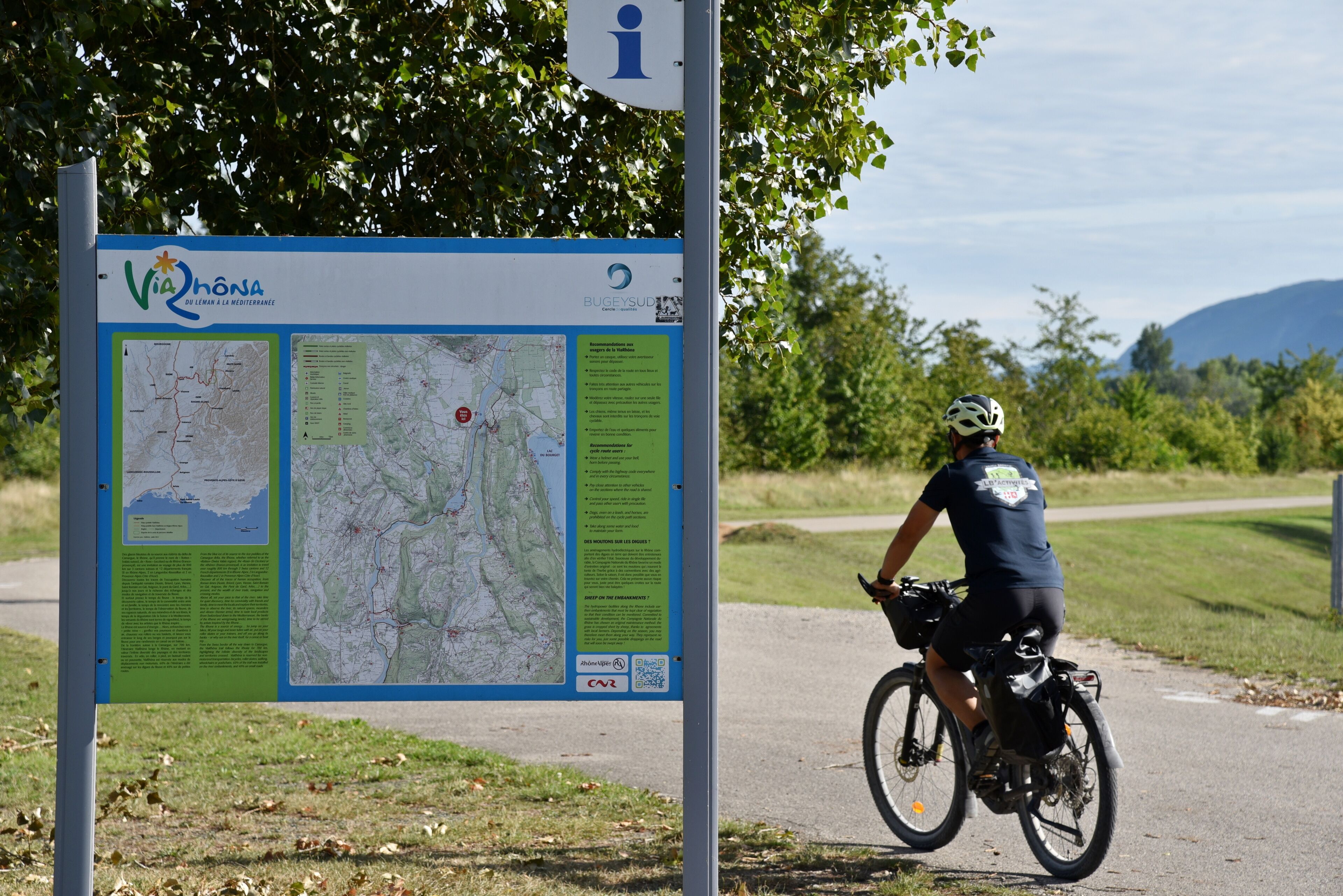 A map sign with text and a QR code, a cyclist on a path, and a natural landscape with trees and hills.