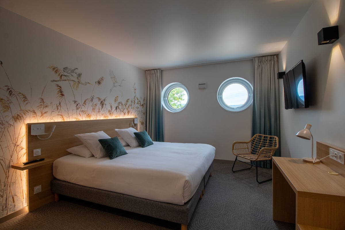 A modern hotel room with a large bed, two windows, a desk, and a chair.