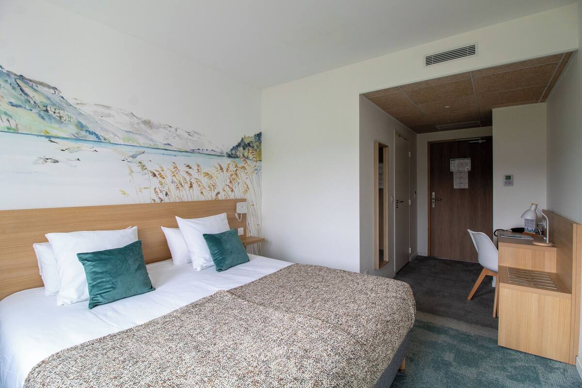 A hotel room with a bed, a desk, and a painting on the wall.