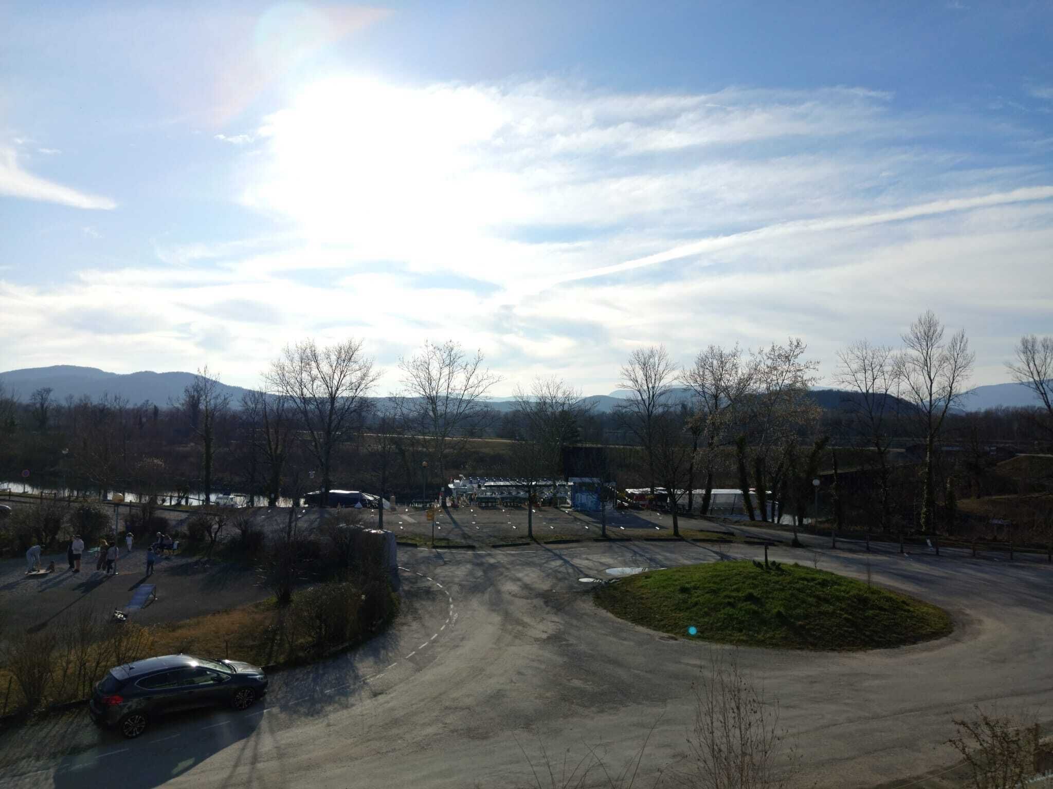 Un espace de stationnement avec quelques voitures, un petit coin de verdure et une vue lointaine sur des montagnes sous un ciel partiellement nuageux.
