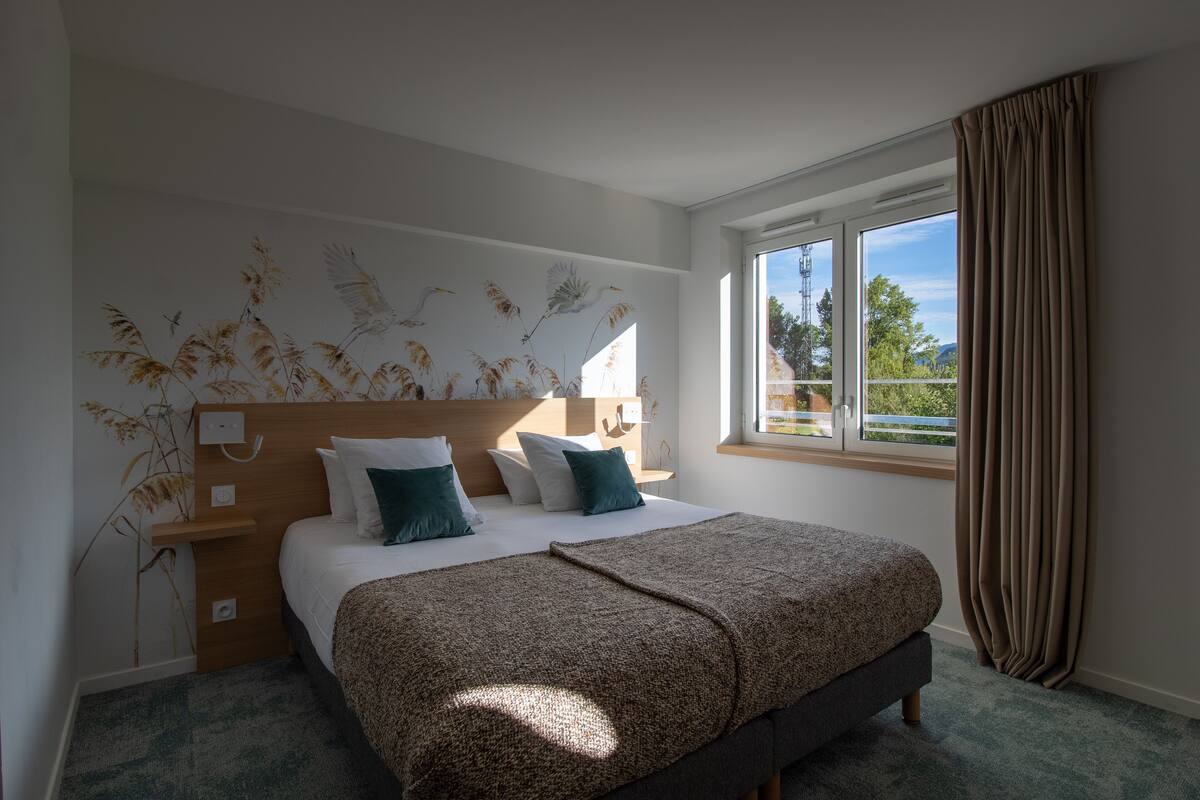 A bedroom with a bed, a nightstand, a window with curtains, and a wall-mounted light fixture.