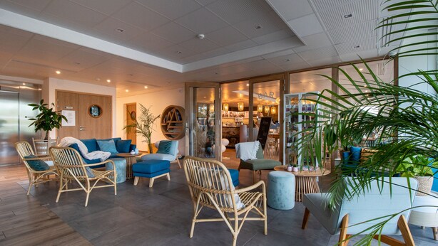 A modern lounge area with wicker furniture, a blue sofa, and a bar visible through the glass doors. -  Shana Hotel & Residence (Chanaz)