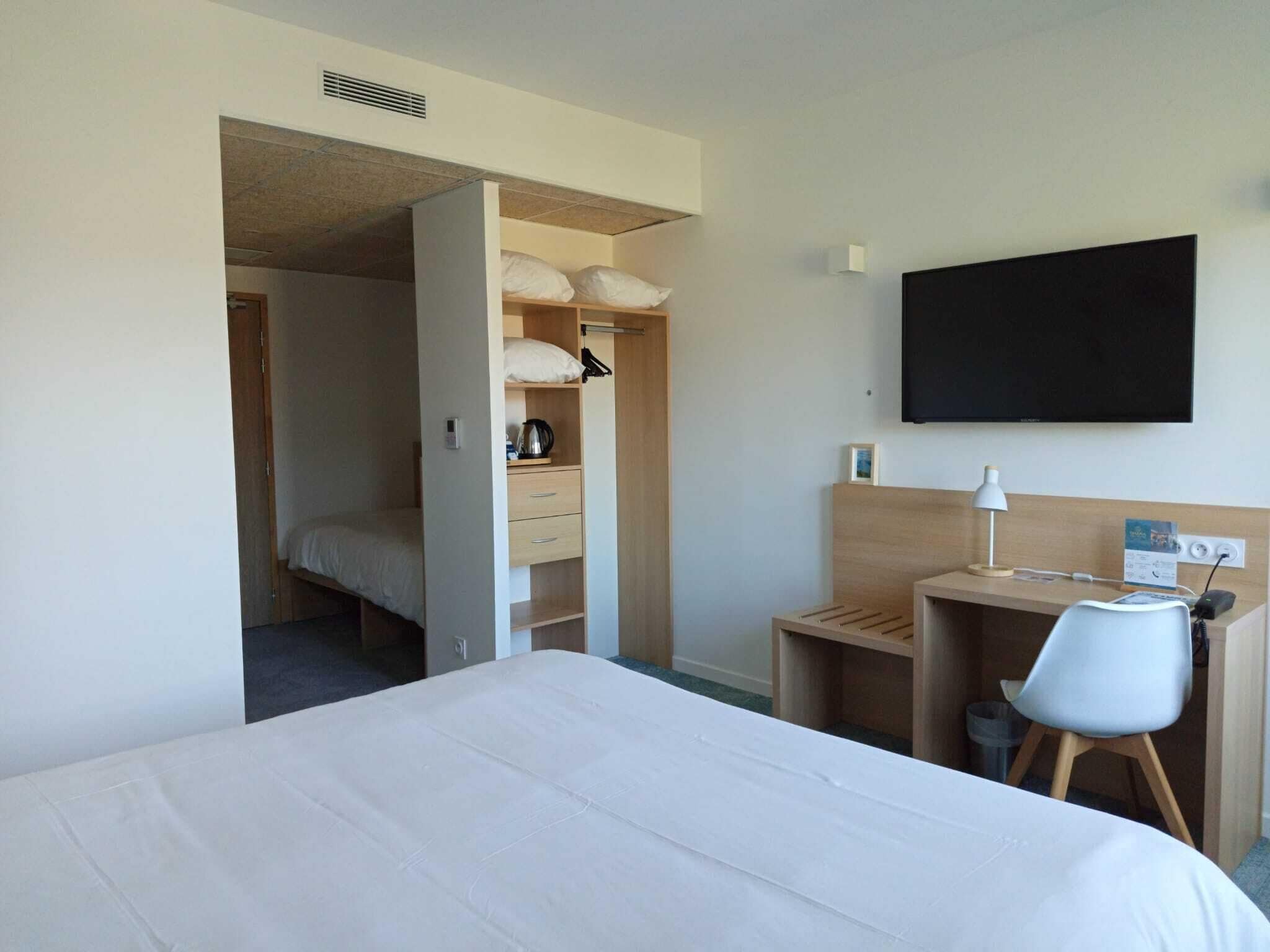 Quarto de hotel com cama, escrivaninha com cadeira, guarda-roupa e televisão de tela plana na parede.