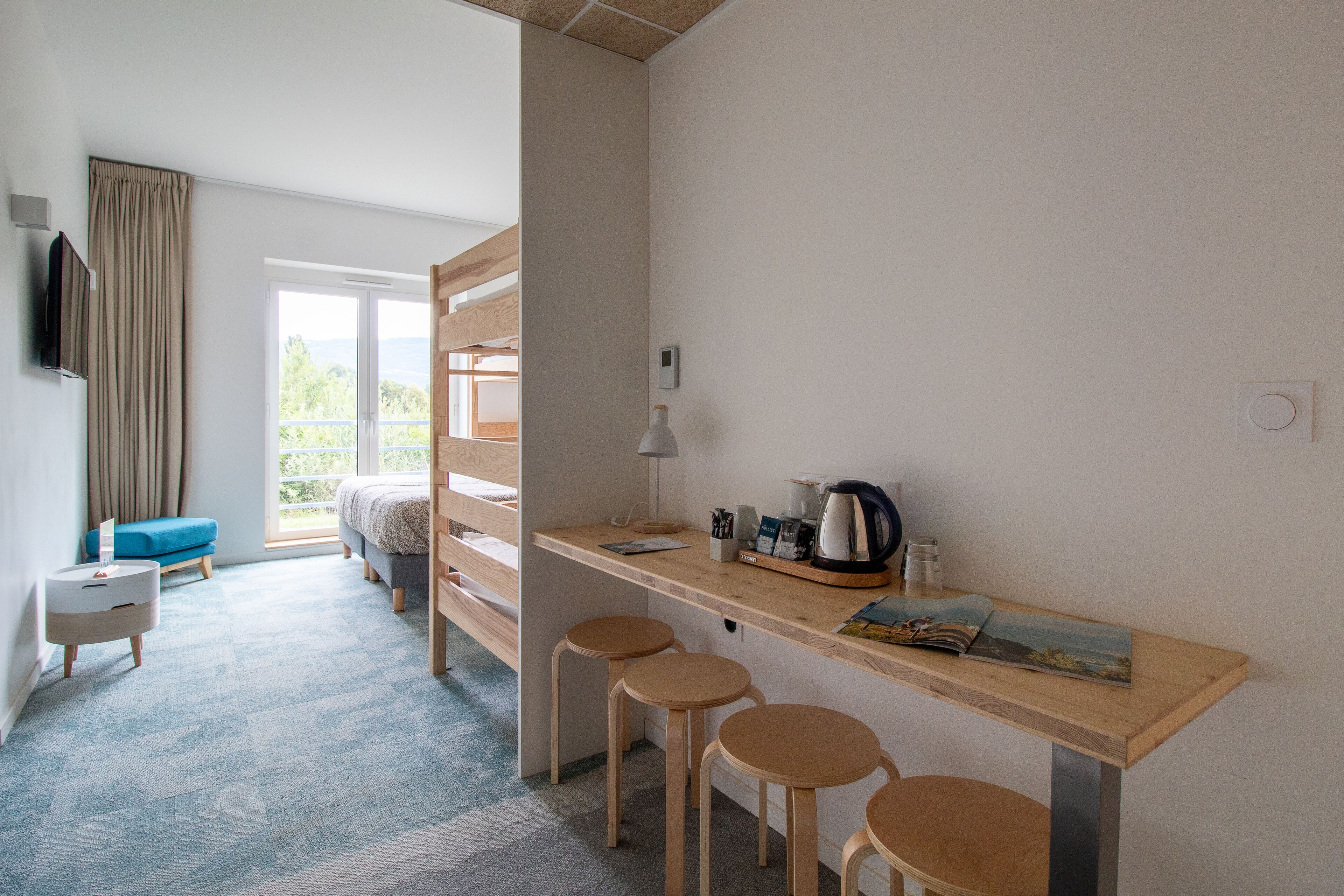A modern hotel room with a wooden desk, two stools, a bed, a TV, and a view of the outdoors.