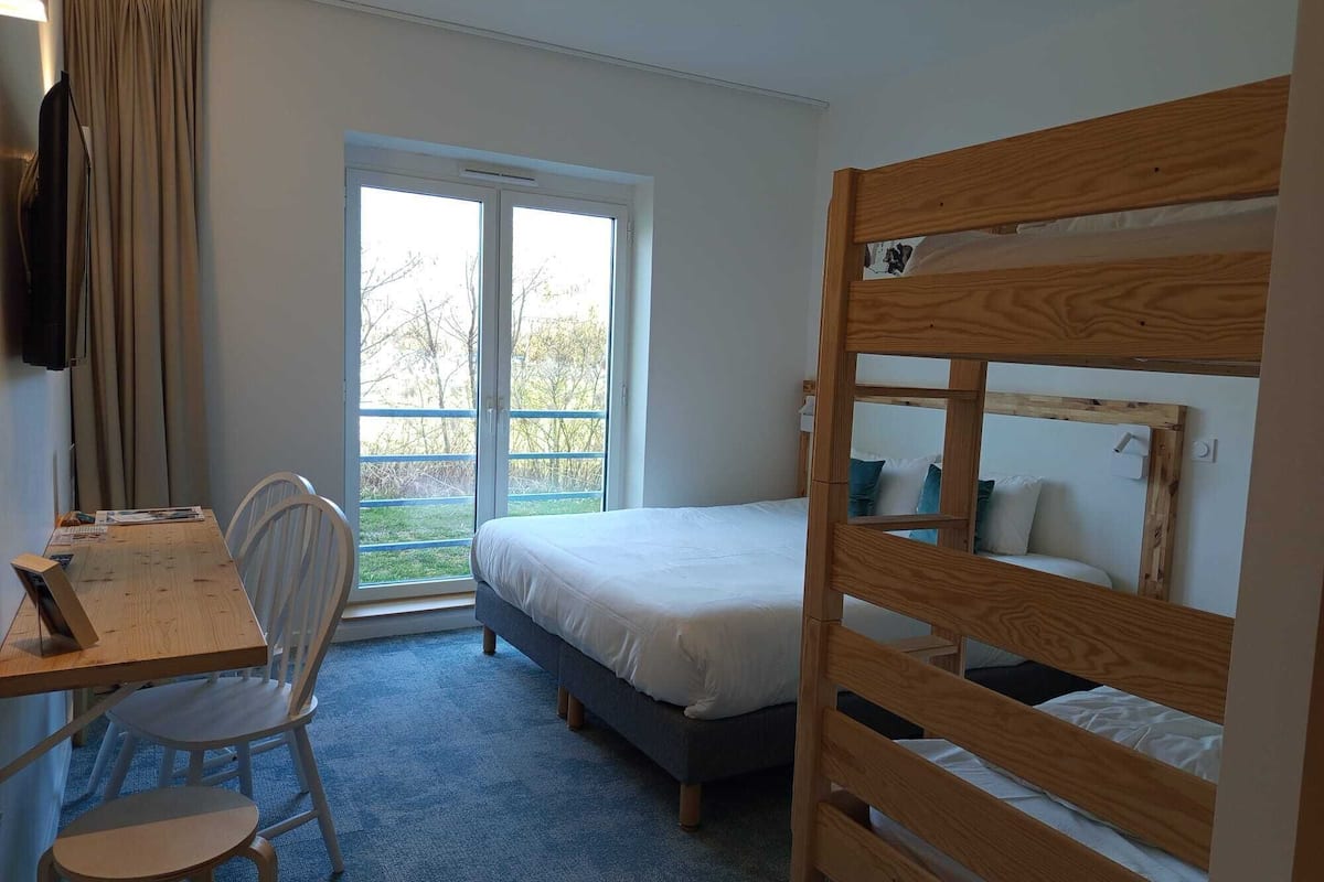 A room with a bunk bed, a desk with a chair, a window with curtains, and a view of the outdoors.