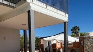 Een terras buiten met witte stoelen en een overdekte zithoek, een stenen muur en uitzicht op een gebouw met een balkon.