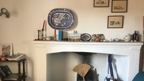 A living room with a fireplace, a blue armchair, a wooden side table with books, and a decorative plate on the mantel. - Apartment (Sant'Ilario)