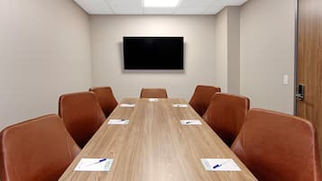 A conference room with a long wooden table, six brown leather chairs, and a flat-screen TV mounted on the wall.