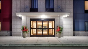 A hotel entrance with a glass door, two potted plants with red flowers, and a lighted sign above the door. - Candlewood Suites Detroit Sterling Heights by IHG (Macomb)