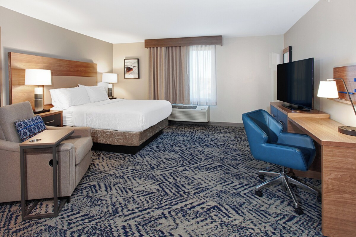 A hotel room with a bed, a desk, a chair, a TV, and an air conditioning unit.
