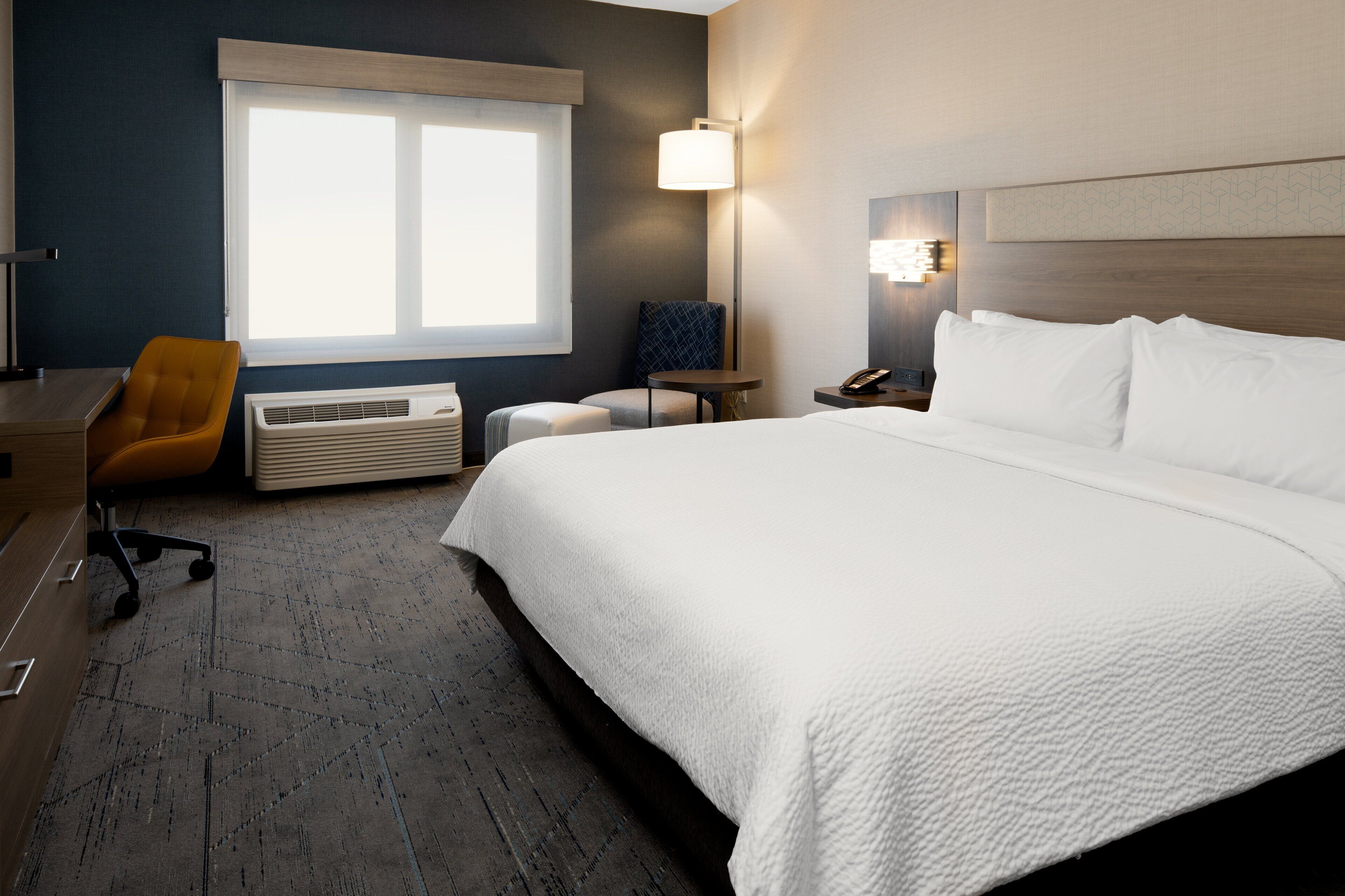 Une chambre d’hôtel avec un grand lit, un bureau avec une chaise, une petite table, une lampe et une fenêtre avec des stores.