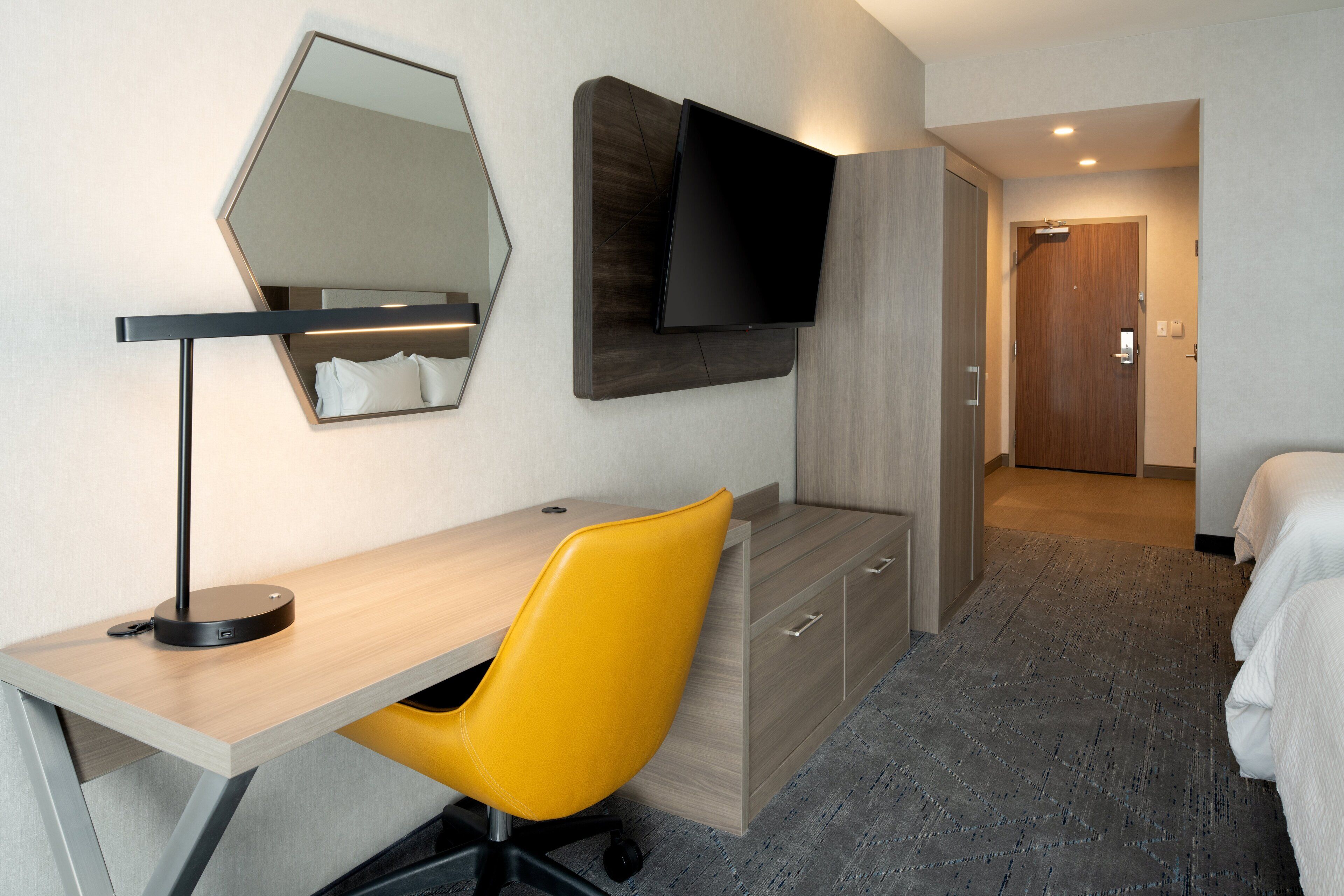 Ein Hotelzimmer mit Schreibtisch, einem gelben Sessel, einem an der Wand befestigten Flachbildfernseher, einem Holzschrank und einem Bett mit weißer Bettwäsche.