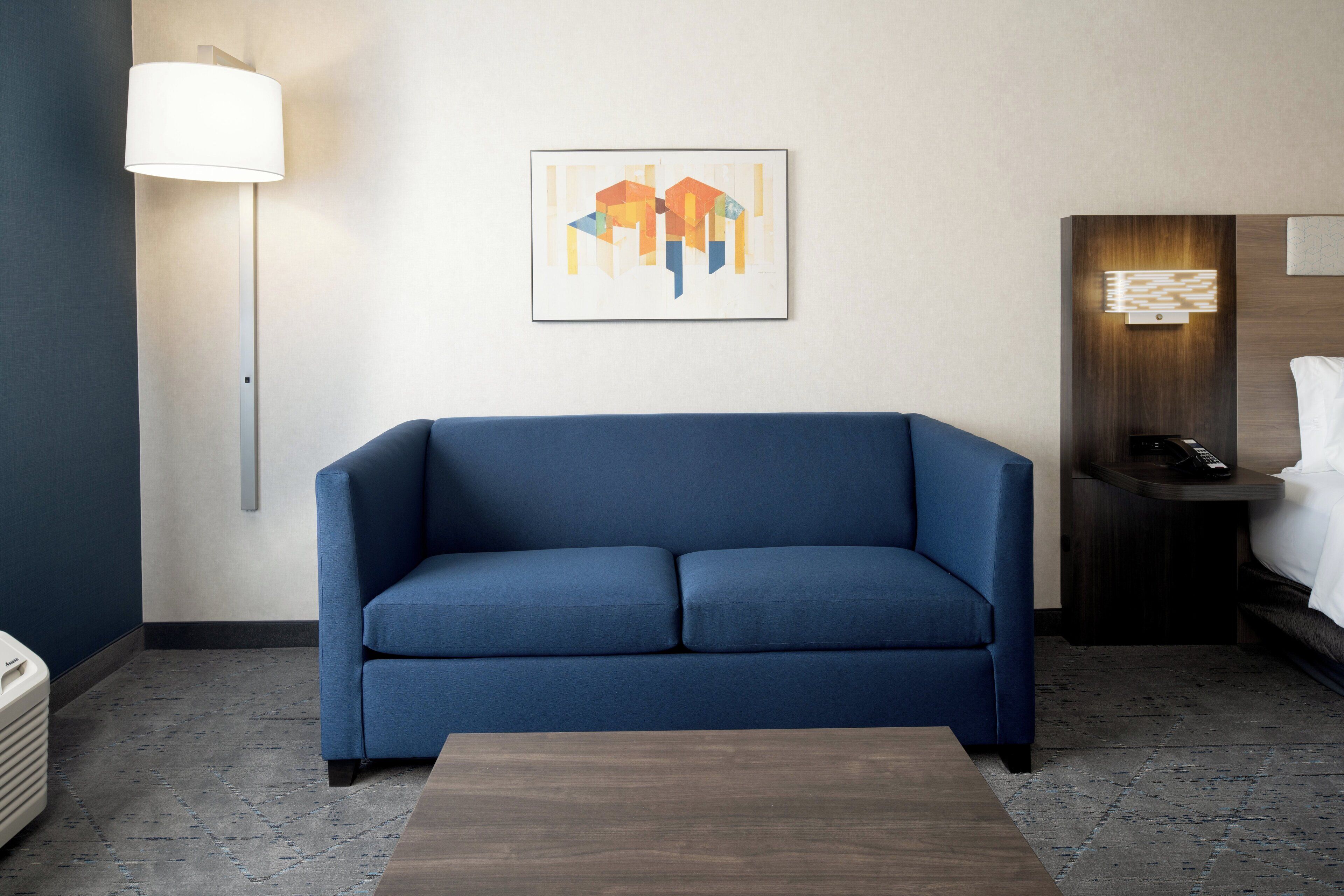 A modern hotel room with a blue sofa, a wooden coffee table, a floor lamp, a painting on the wall, a bedside table with a lamp, and a bed with white linens.