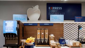 Uma estação de café da manhã com máquina de café, dispensadores de frutas e cereais, e uma tigela de frutas.