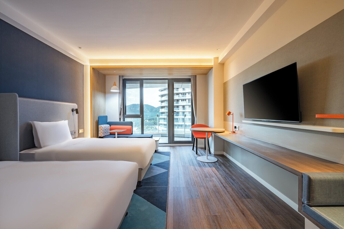 A modern hotel room with two beds, a TV, a desk, and a view of a building through the window.